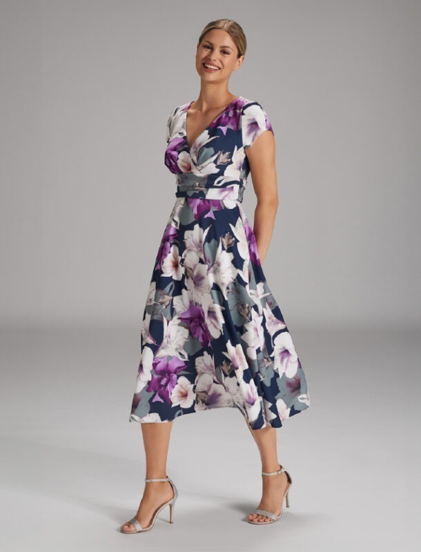 Eine Frau trägt ein blumiges Kleid in Blau- und Lilatönen und steht lächelnd mit elegantem Schuhwerk.