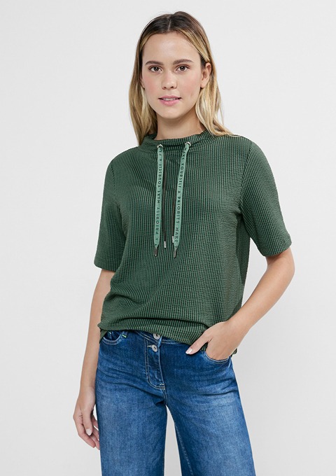 Eine Person trägt ein grünes, kurzärmliges Shirt mit einer Kapuze und Bindebändern, kombiniert mit blauen Jeans.