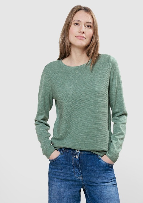 Eine Frau trägt einen grünen Pulli mit langem Arm und steht mit den Händen in den Taschen ihrer Jeans.