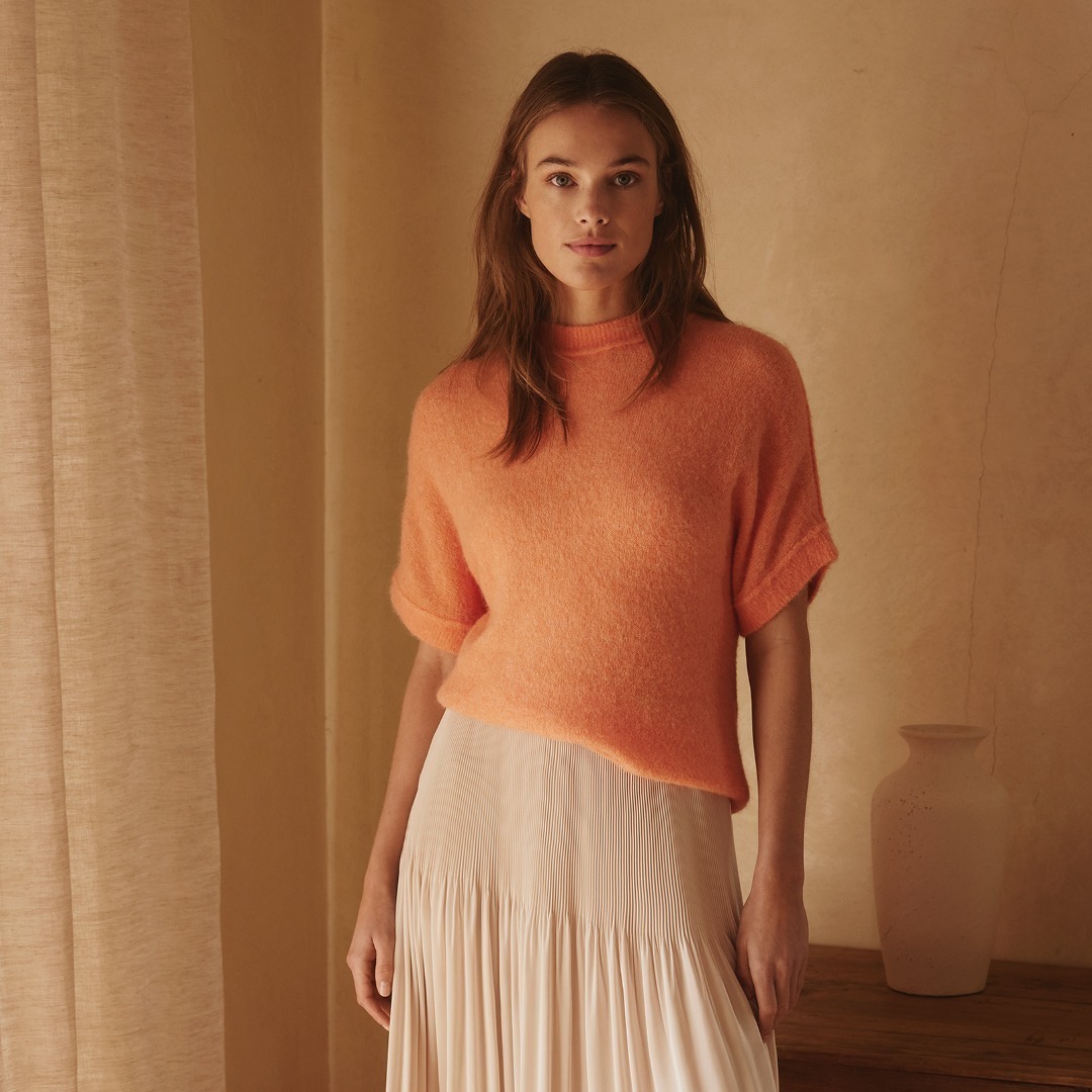 Eine Frau trägt einen orangefarbenen Pullover und einen langen, plissierten Rock. Sie steht vor einem neutralen Hintergrund mit sanftem Licht.