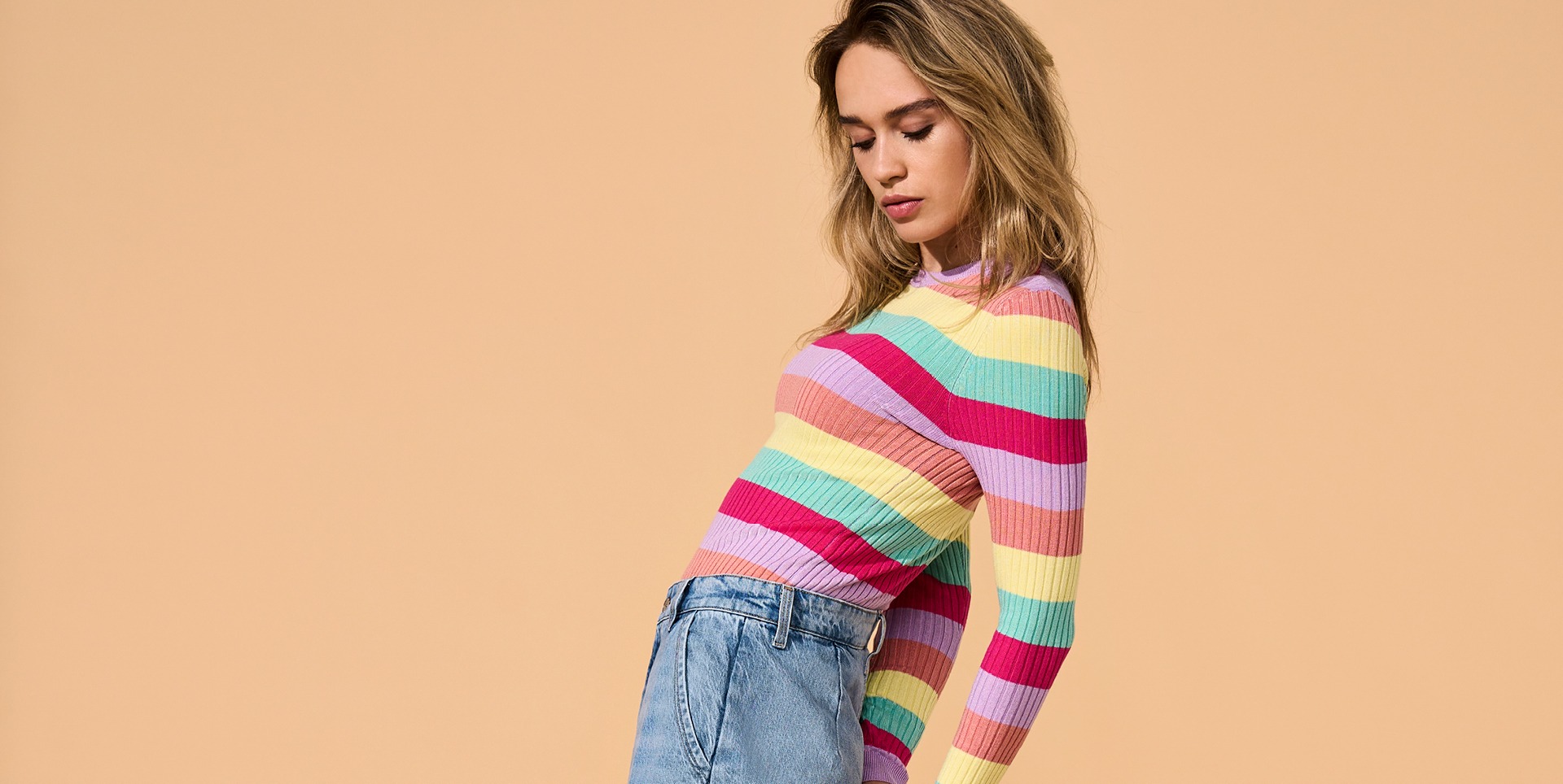 Ein Modell trägt einen gestreiften Pullover in bunten Farben und eine Jeans, vor einem einfarbigen Hintergrund.