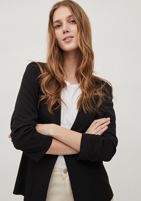 Eine Frau mit langen, lockigen Haaren trägt einen schwarzen Blazer und ein weißes Oberteil. Sie steht mit verschränkten Armen und lächelt.