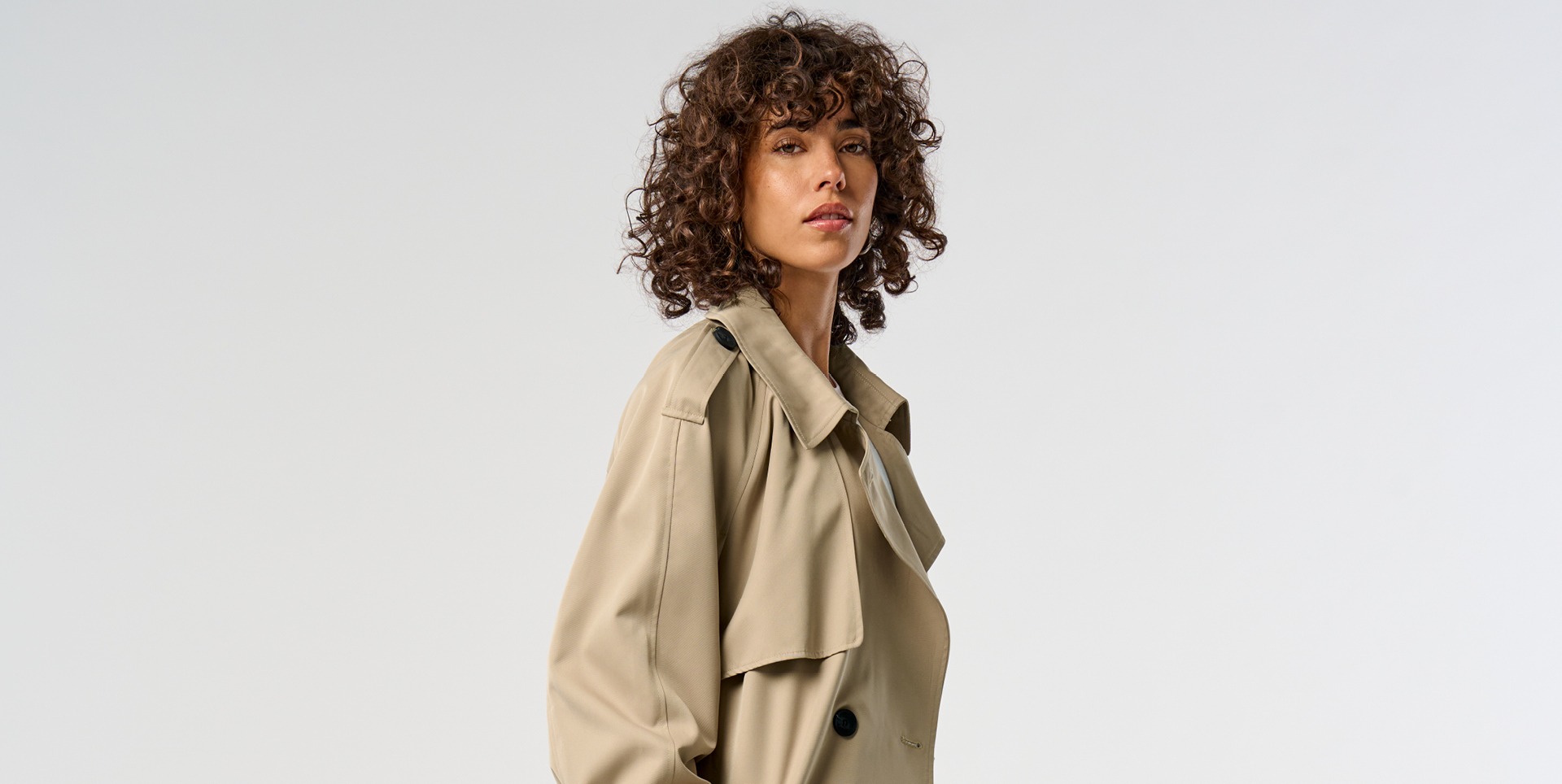 Eine Frau mit lockigem Haar trägt einen beigen Trenchcoat. Eine Frau mit lockigem Haar trägt einen beigen Trenchcoat.