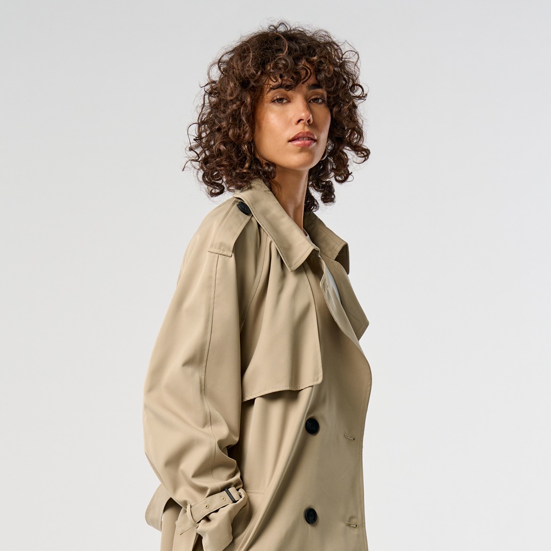 Eine Frau mit lockigem Haar trägt einen beigen Trenchcoat und blickt zur Seite. Eine Frau mit lockigem Haar trägt einen beigen Trenchcoat und blickt zur Seite.