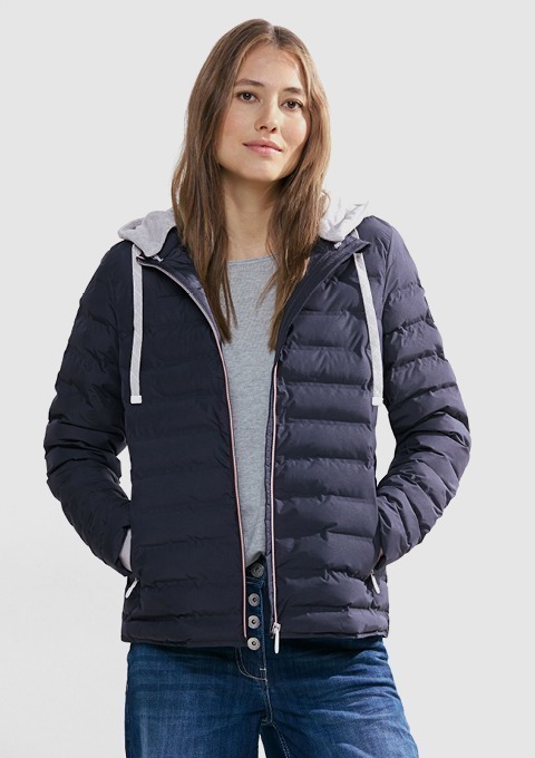 Eine Frau trägt eine dunkelblaue, gepolsterte Jacke mit Kapuze und grauem T-Shirt, dazu eine Jeans. Eine Frau trägt eine dunkelblaue, gepolsterte Jacke mit Kapuze und grauem T-Shirt, dazu eine Jeans.