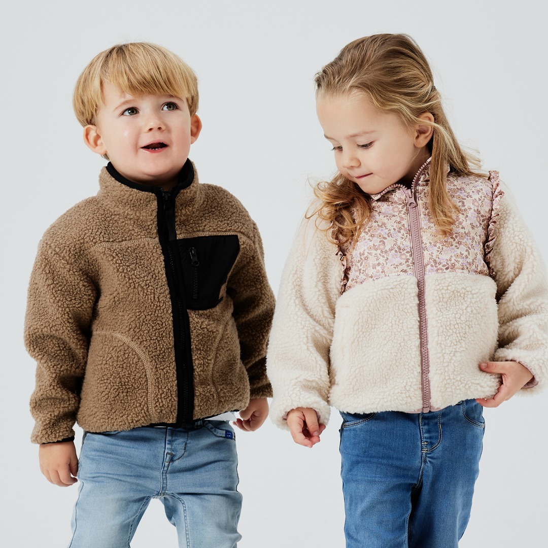 Zwei Kinder in stylischen Fleecejacken stehen nebeneinander. Das Mädchen trägt eine helle Jacke mit floralem Muster, der Junge eine braune Jacke mit schwarzem Reißverschluss. Zwei Kinder in stylischen Fleecejacken stehen nebeneinander. Das Mädchen trägt eine helle Jacke mit floralem Muster, der Junge eine braune Jacke mit schwarzem Reißverschluss.