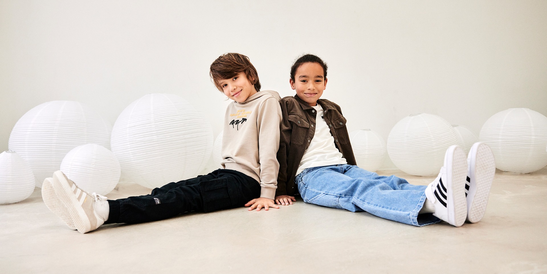 Zwei Jungen sitzen auf dem Boden, umgeben von weißen Papierlaternen. Der Junge links trägt einen hellen Hoodie und schwarze Hose, der rechte einen braunen Oberteil und blaue Jeans.
