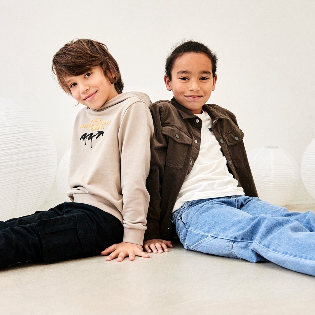 Zwei Kinder sitzen nebeneinander auf dem Boden und lächeln. Sie tragen lässige Oberbekleidung.