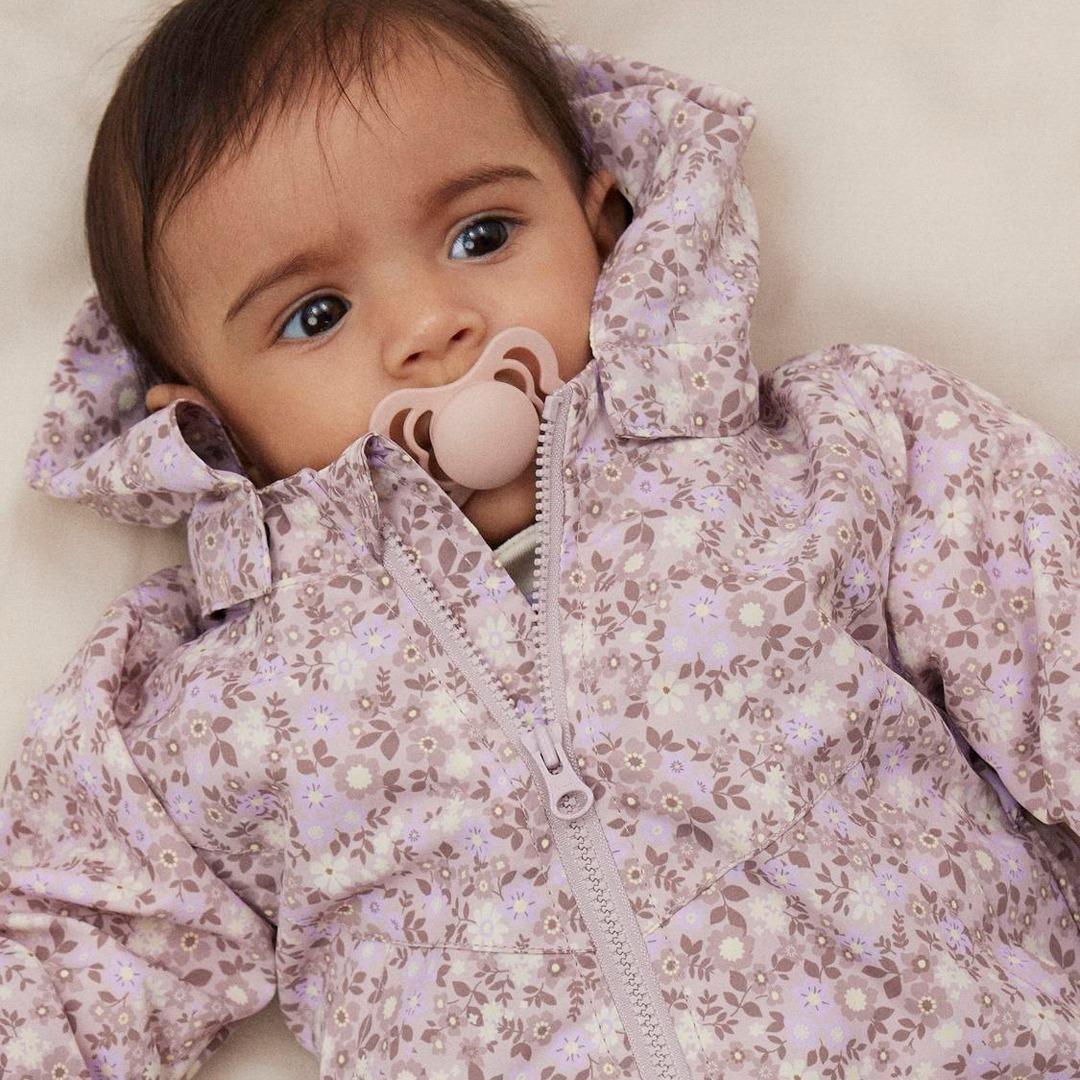 Ein Baby mit einem Schnuller trägt eine lila gemusterte Jacke.