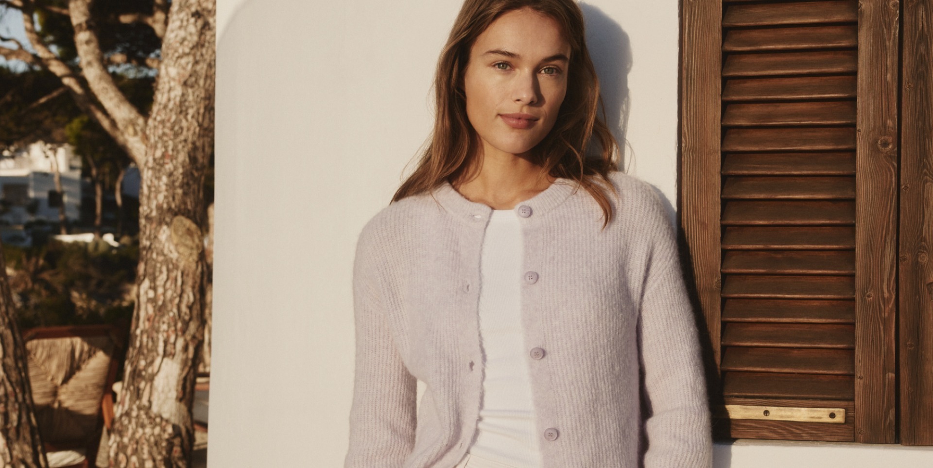 Junge Frau in einem lila Strickcardigan vor einer weißen Wand und Holzjalousien. Junge Frau in einem lila Strickcardigan vor einer weißen Wand und Holzjalousien.