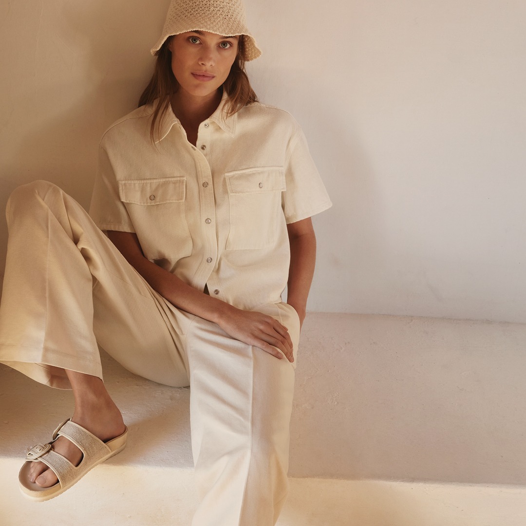 Eine Person sitzt in einem beigen Outfit mit einem Hut. Der Hintergrund ist hell und neutral gehalten. Eine Person sitzt in einem beigen Outfit mit einem Hut. Der Hintergrund ist hell und neutral gehalten.