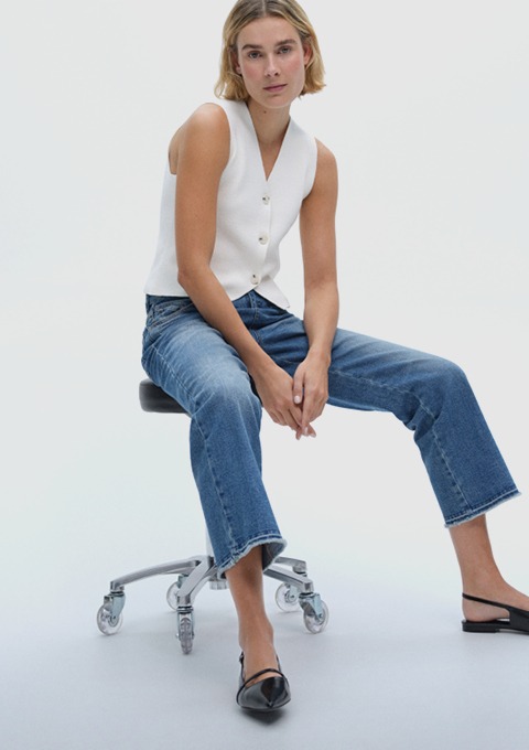 Eine Frau sitzt auf einem Drehstuhl und trägt einen weißen ärmellosen Pullover sowie Jeans. Sie trägt schwarze Sandalen und Blick zur Kamera. Eine Frau sitzt auf einem Drehstuhl und trägt einen weißen ärmellosen Pullover sowie Jeans. Sie trägt schwarze Sandalen und Blick zur Kamera.