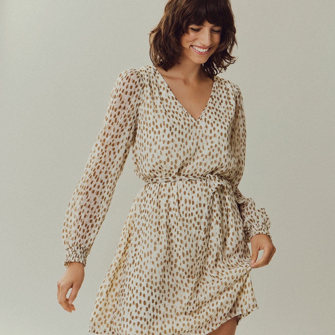 Eine lächelnde Frau trägt ein luftiges Kleid mit einem Punktmuster in beige und braun.