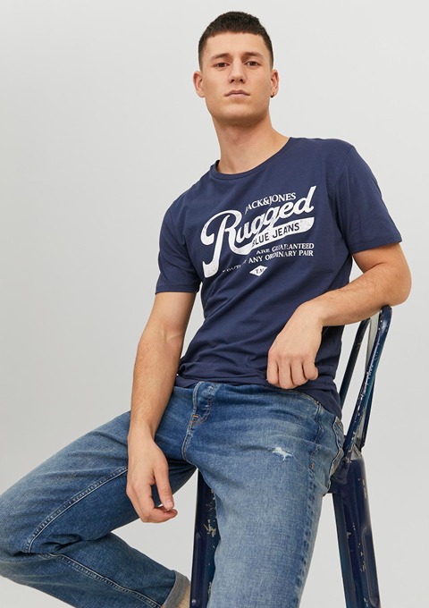 Ein junger Mann sitzt auf einem blauen Stuhl und trägt ein dunkelblaues T-Shirt mit dem Schriftzug "Rugged" und Jeans.