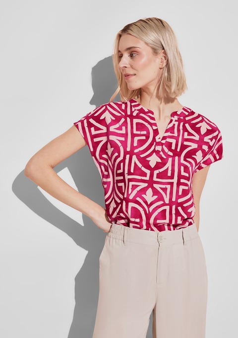 Eine Frau trägt ein pinkes Oberteil mit einem geometrischen Muster und beigen Hosen.