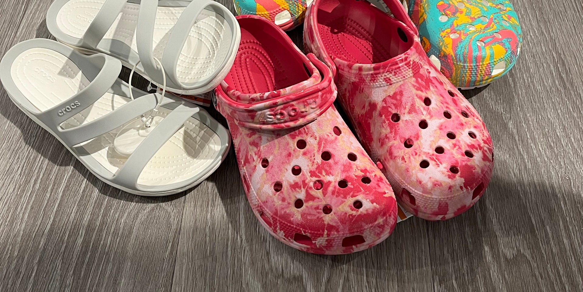 Drei Paar Schuhe: rosa Crocs, weiße Sandalen und ein buntes Paar.