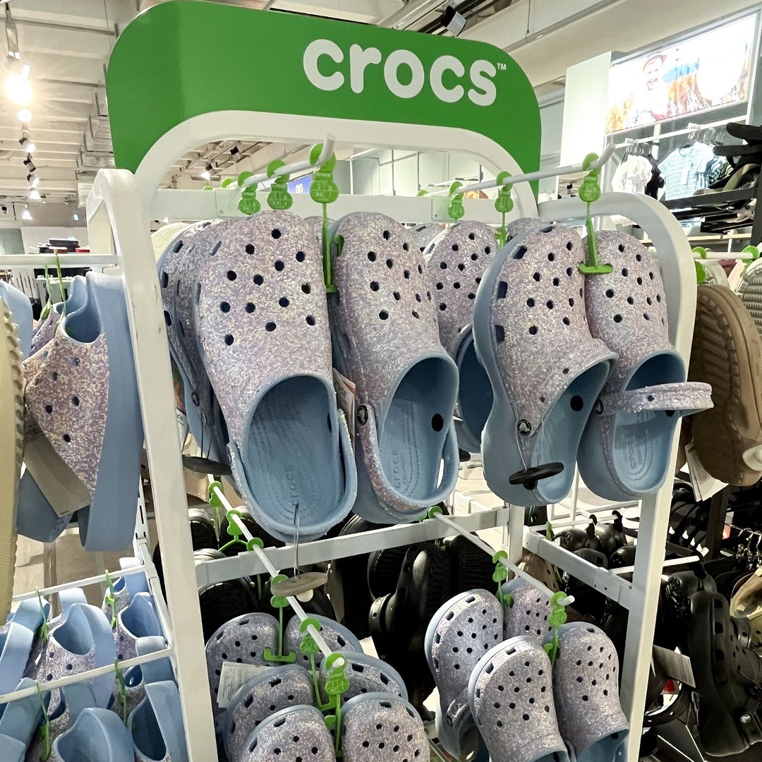 Eine Anzeige von Crocs in einer Verkaufsstelle mit lila-glitzernden Clogs auf einem Kleidungsständer.
