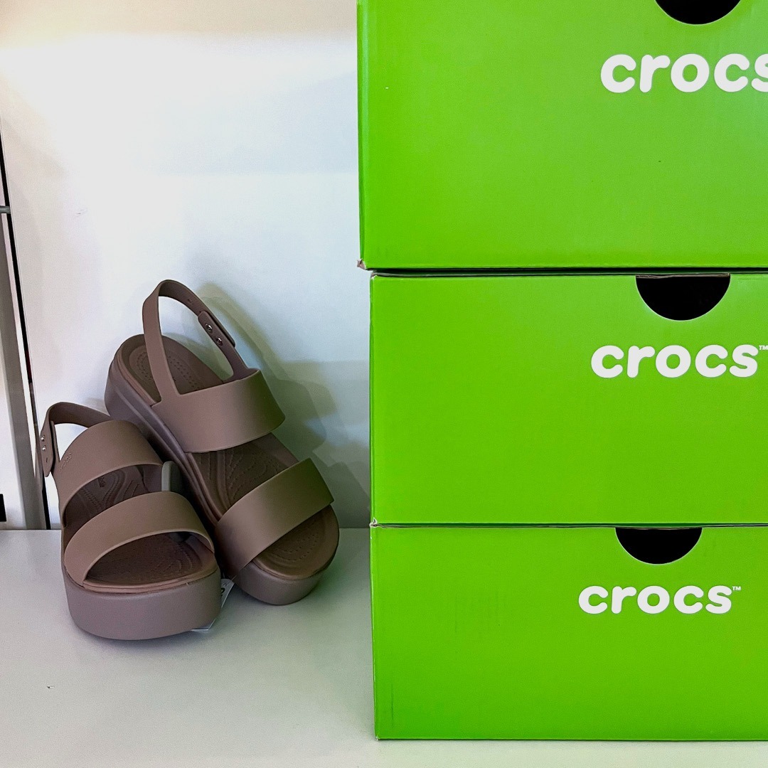 Ein Paar braune Crocs-Sandalen neben grünen Crocs-Verpackungen.
