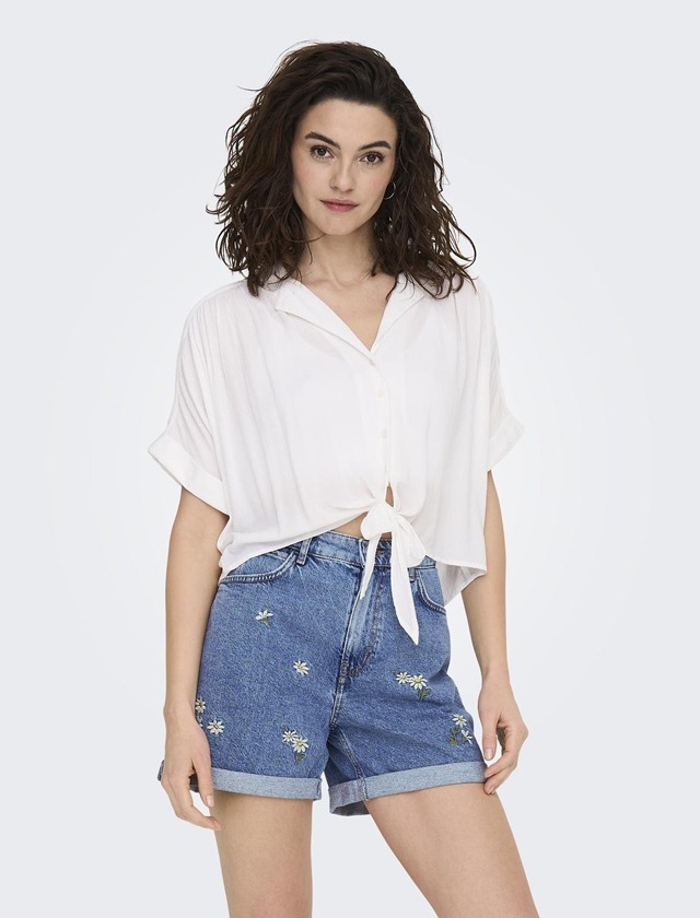 Eine Person trägt ein weißes, lässiges Hemd und blaue Jeansshorts mit Blumenstickerei.