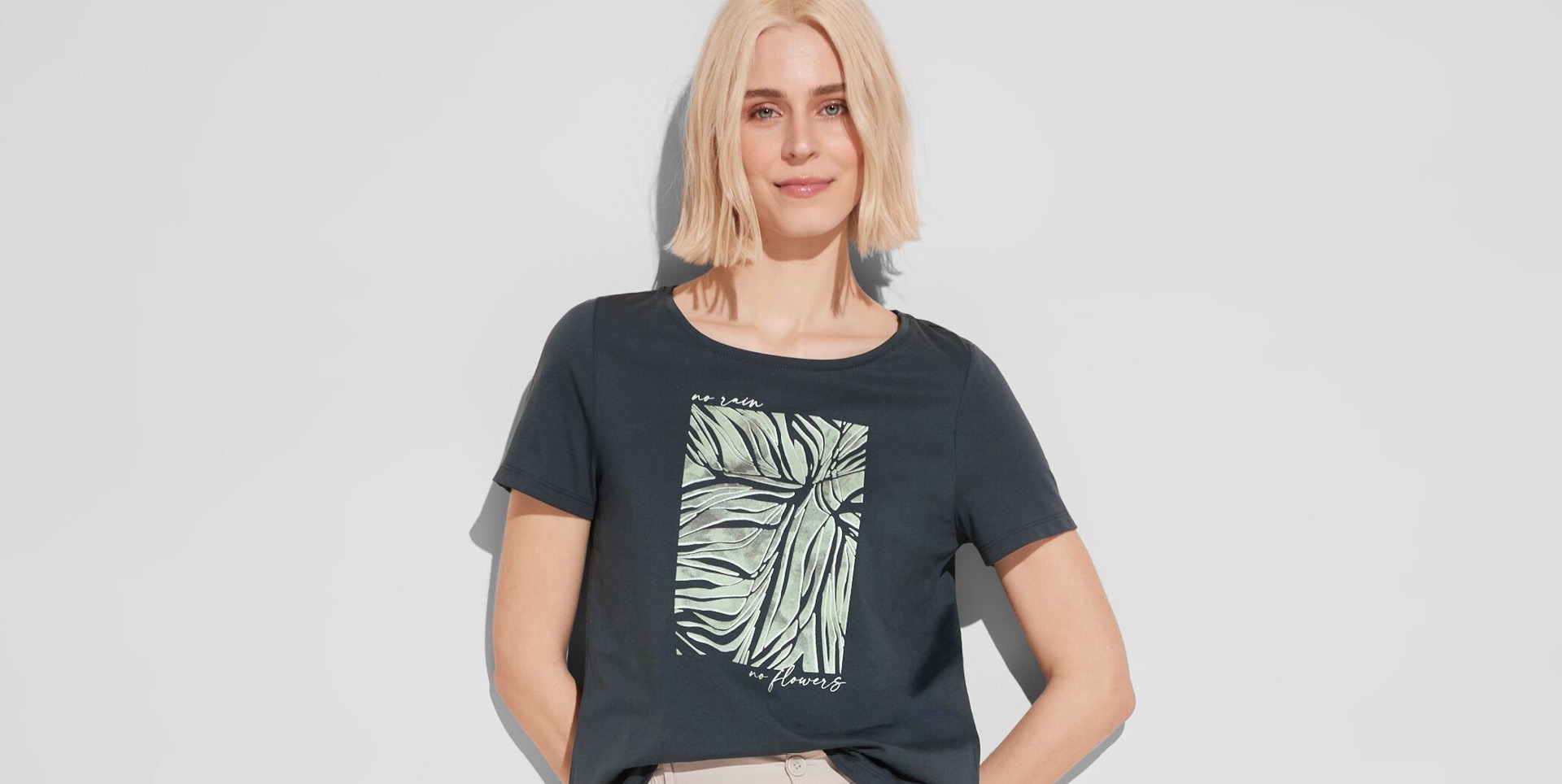 Eine Frau trägt ein schwarzes T-Shirt mit einem Blattmuster und dem Text "no rain no flowers".