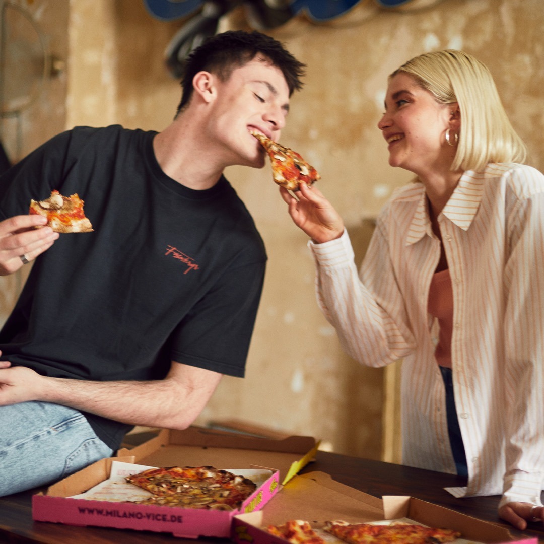 Zwei Personen sitzen am Tisch und essen Pizza. Sie teilen einen fröhlichen Moment, während sie jeweils ein Stück Pizza greifen. Im Hintergrund sind Pizzakartons zu sehen.