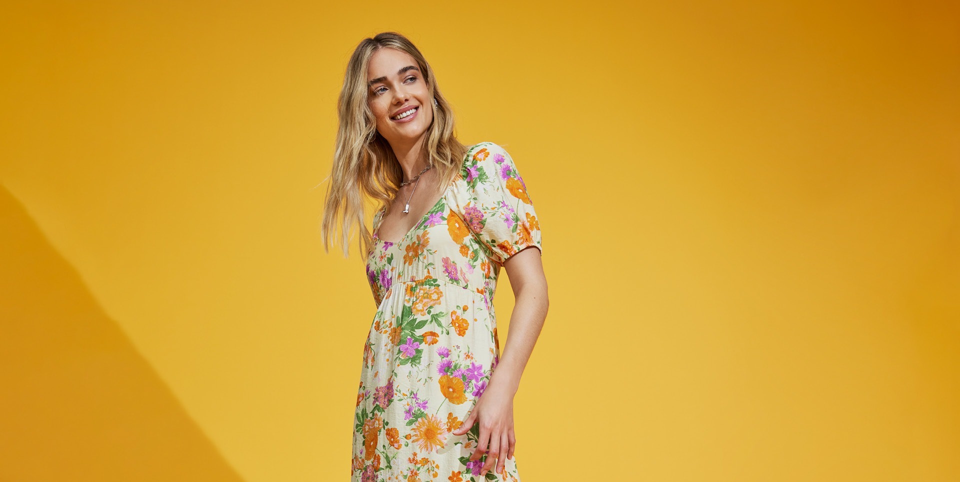 Eine lächelnde Frau in einem blumigen Kleid vor einem gelben Hintergrund.