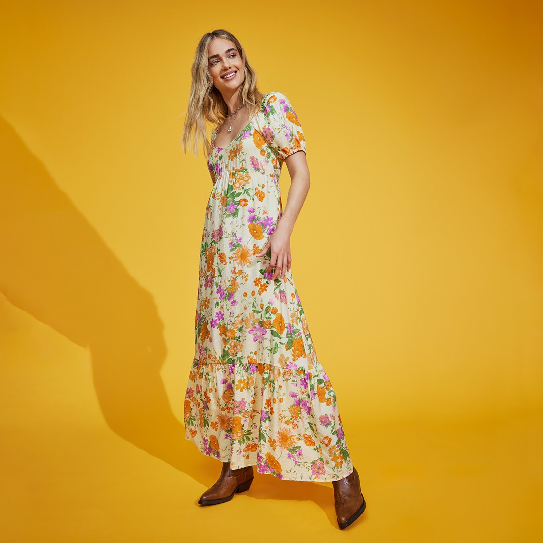 Eine Frau trägt ein blumiges Maxikleid vor einem gelben Hintergrund.
