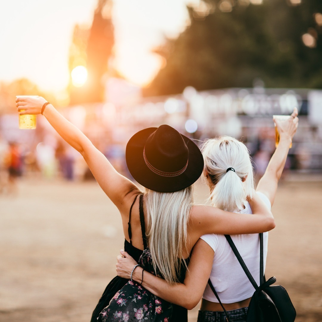 Zwei Frauen umarmen sich und heben ihre Gläser in die Luft. Sie stehen vor einer Festivalkulisse bei Sonnenuntergang.