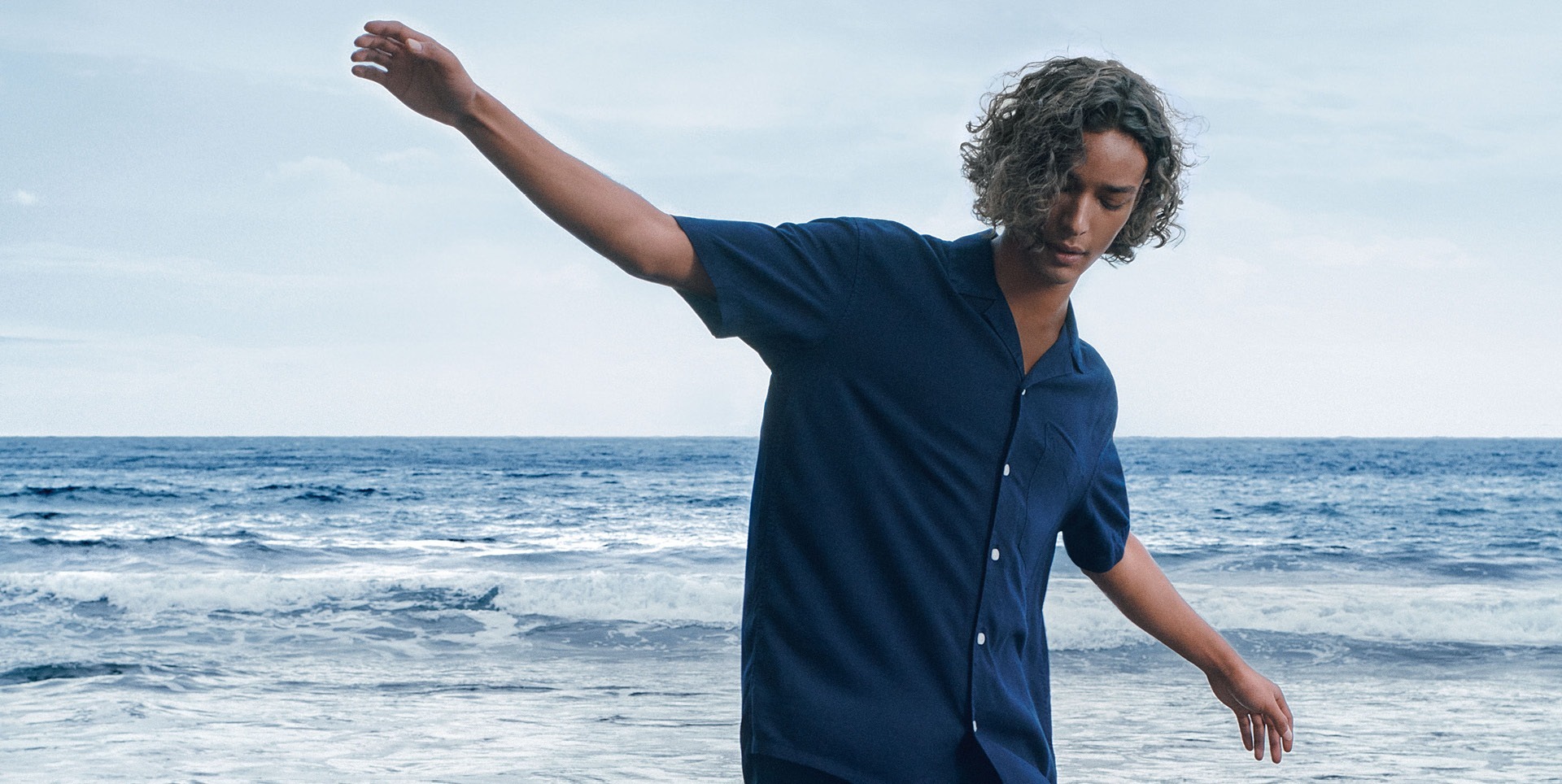 Ein junger Mann mit lockigem Haar steht am Meer und schaut nach unten. Ein junger Mann mit lockigem Haar steht am Meer und schaut nach unten.