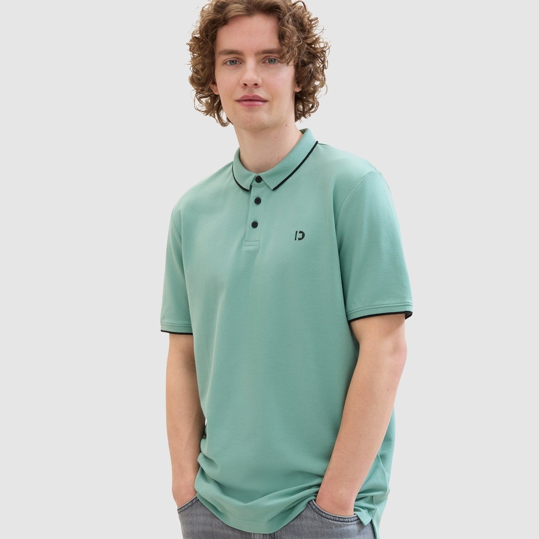 Ein junger Mann mit lockigem Haar trägt ein mintgrünes Poloshirt mit schwarzem Kragen und einer kleinen Logo-Stickerei. Ein junger Mann mit lockigem Haar trägt ein mintgrünes Poloshirt mit schwarzem Kragen und einer kleinen Logo-Stickerei.