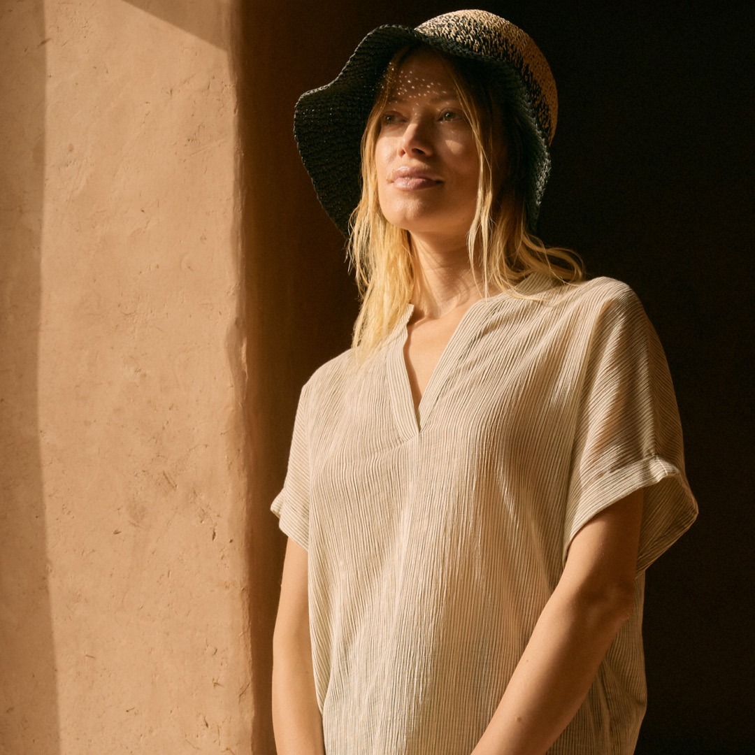 Eine blonde Frau trägt ein lockeres, gestreiftes Oberteil und einen großen Sonnenhut. Sie steht vor einer wand und lächelt. Eine blonde Frau trägt ein lockeres, gestreiftes Oberteil und einen großen Sonnenhut. Sie steht vor einer wand und lächelt.