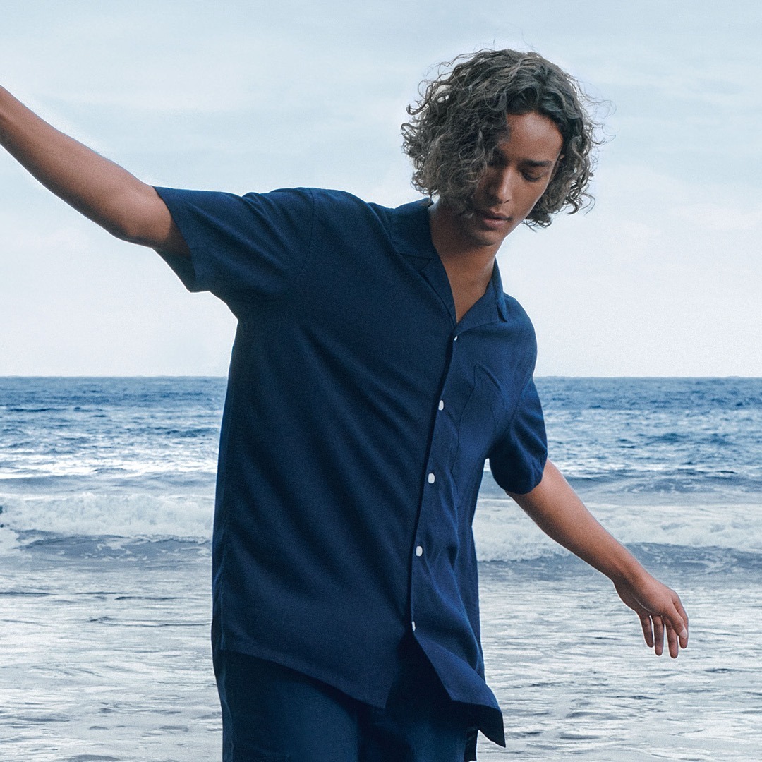 Ein junger Mann mit lockigem Haar steht am Strand und schaut nach unten, während er einen Arm hebt. Im Hintergrund ist das Meer sichtbar.