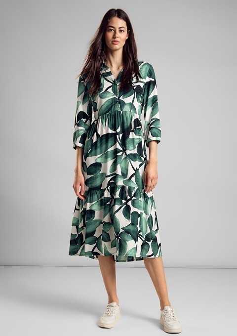 Eine Frau trägt ein langes, blumenmotiviertes Kleid mit grünem Blattmuster und weißen Sneakers. Eine Frau trägt ein langes, blumenmotiviertes Kleid mit grünem Blattmuster und weißen Sneakers.