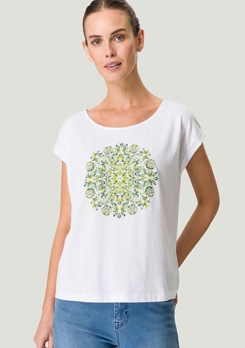Eine Frau trägt ein weißes T-Shirt mit einem bunten, mandalaartigen Muster in Grün- und Blautönen. Eine Frau trägt ein weißes T-Shirt mit einem bunten, mandalaartigen Muster in Grün- und Blautönen.
