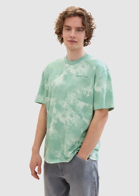 Ein junger Mann trägt ein grünes T-Shirt mit Muster und steht lächelnd vor einem neutralen Hintergrund. Ein junger Mann trägt ein grünes T-Shirt mit Muster und steht lächelnd vor einem neutralen Hintergrund.