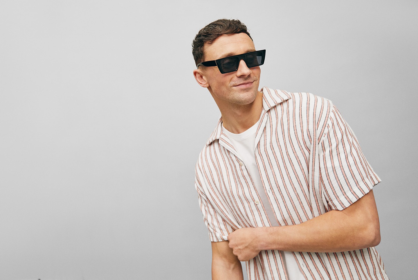Ein lächelnder Mann trägt eine gestreifte Hemdbluse und Sonnenbrillen vor einem grauen Hintergrund.
