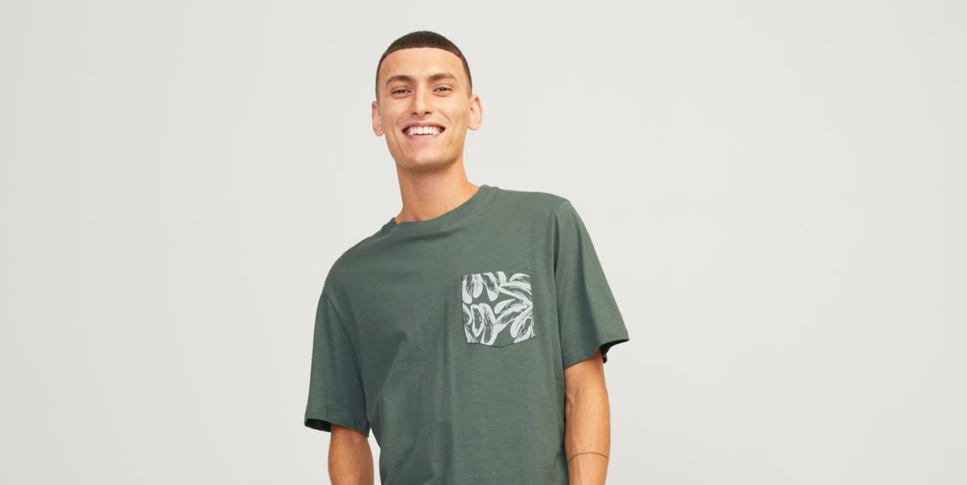 Ein Mann mit kurzem Haar trägt ein dunkelgrünes T-Shirt mit einer grauen, gemusterten Brusttasche und lächelt. Ein Mann mit kurzem Haar trägt ein dunkelgrünes T-Shirt mit einer grauen, gemusterten Brusttasche und lächelt.