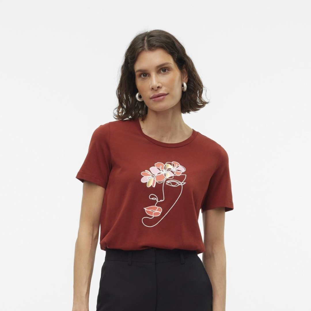 Eine Frau trägt ein rotbraunes T-Shirt mit einer künstlerischen Zeichnung eines Gesichts und Blumen.