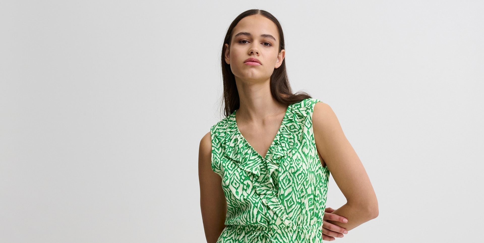Eine junge Frau trägt ein ärmelloses, grßnes Kleid mit einem geometrischen Muster und einem seitlichen Schlitz. Eine junge Frau trägt ein ärmelloses, grßnes Kleid mit einem geometrischen Muster und einem seitlichen Schlitz.
