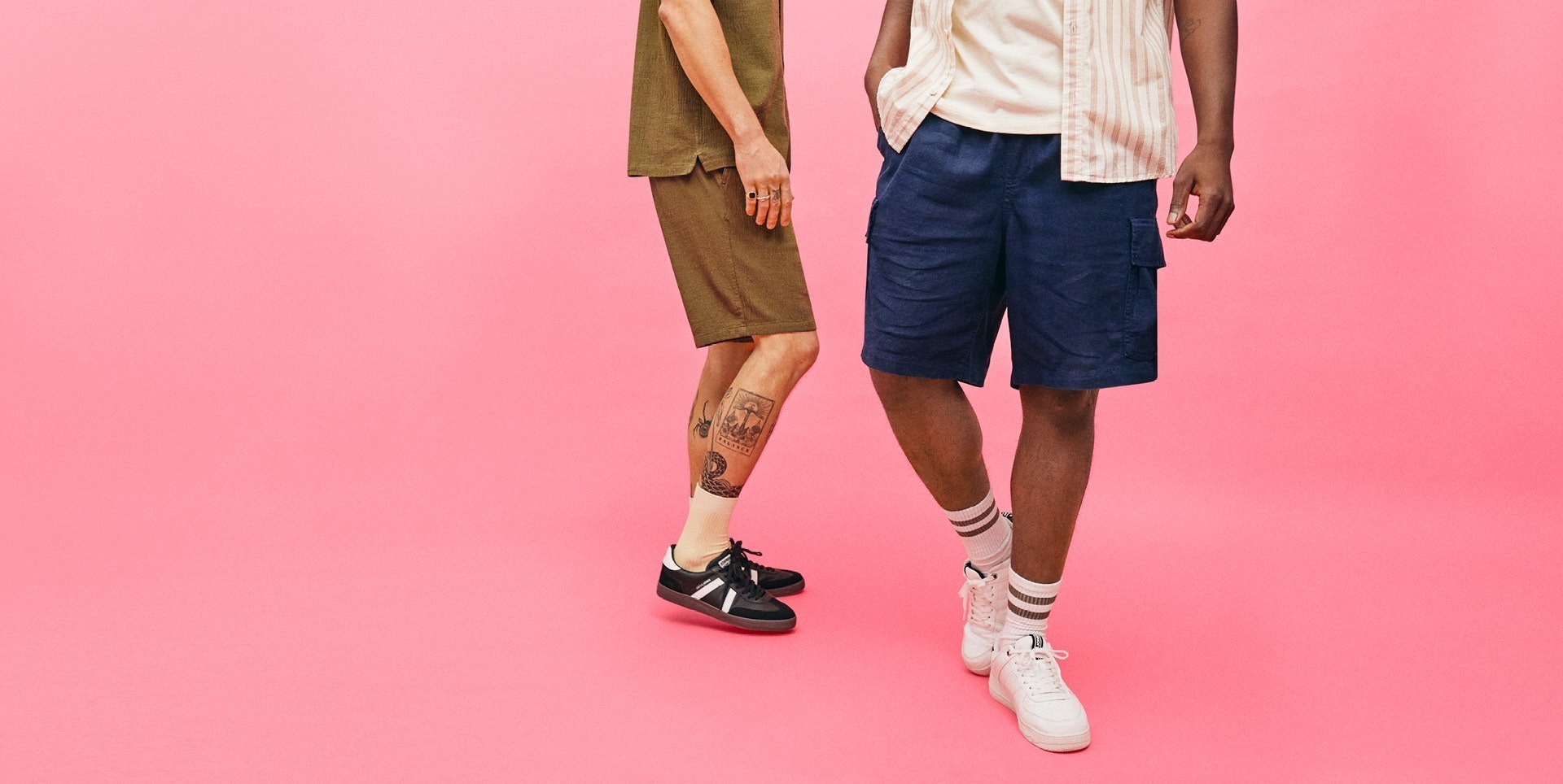 Zwei Personen tragen lässige Kleidung, vor einem pinken Hintergrund. Der eine trägt ein olivfarbenes Oberteil und kurze Hosen, der andere ein gestreiftes Hemd und dunkle Shorts.