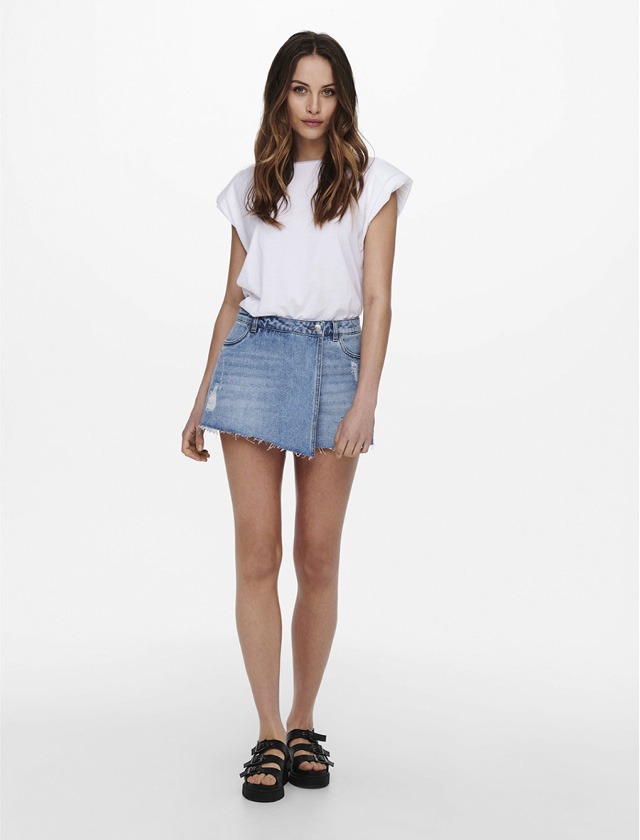 Eine Person steht in einem weißen T-Shirt, kombiniert mit einem Jeansrock und schwarzen Sandalen.
