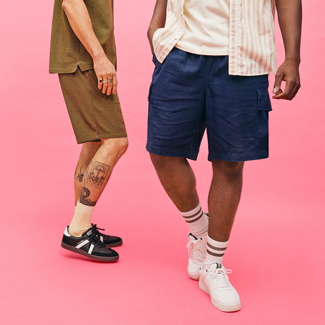 Zwei Männer stehen auf einem pinken Hintergrund, einer trägt eine olivgrüne Hose und schwarze Schuhe, der andere eine blaue Shorts und weiße Sneakers.