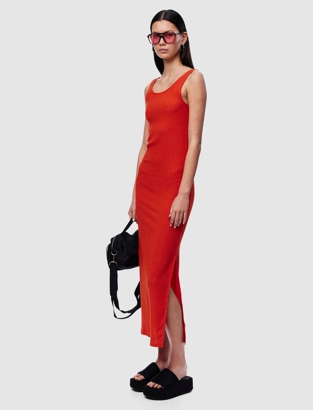 Eine Frau trägt ein enges rotes Kleid, schwarze Sandalen und eine schwarze Tasche. Sie hat lange, glatte Haare und trägt große Sonnenbrillen.