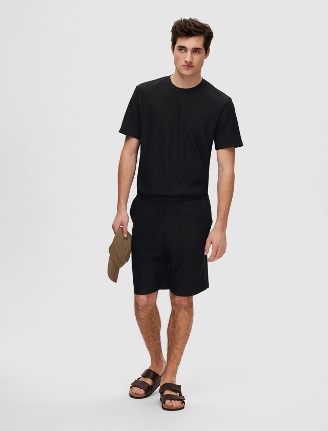 Ein junger Mann trägt ein schwarzes T-Shirt und schwarze Shorts, hält eine beigefarbene Mütze in der Hand und trägt Sandalen. Ein junger Mann trägt ein schwarzes T-Shirt und schwarze Shorts, hält eine beigefarbene Mütze in der Hand und trägt Sandalen.
