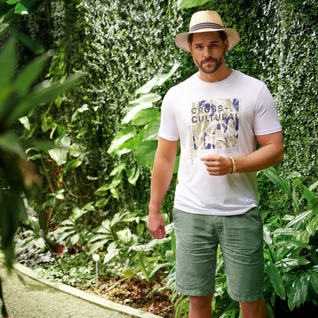 Ein Mann steht in einem grünen, tropischen Garten. Er trägt ein weißes T-Shirt mit Aufdruck und grüne Shorts, dazu einen Strohhut.