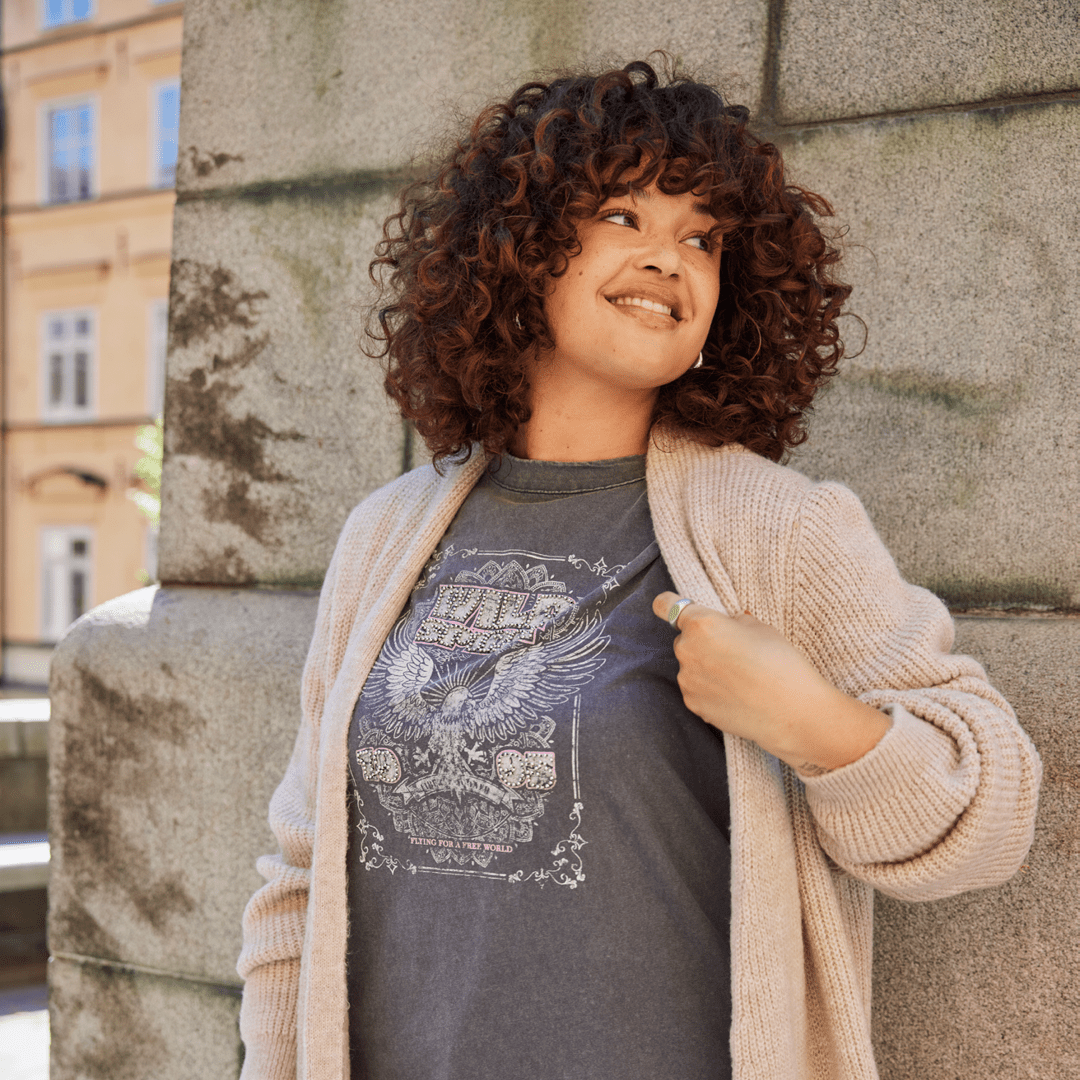Eine lächelnde Frau mit lockigem Haar trägt ein graues T-Shirt und eine helle Strickjacke. Sie steht neben einer Wand und schaut zur Seite. Eine lächelnde Frau mit lockigem Haar trägt ein graues T-Shirt und eine helle Strickjacke. Sie steht neben einer Wand und schaut zur Seite.