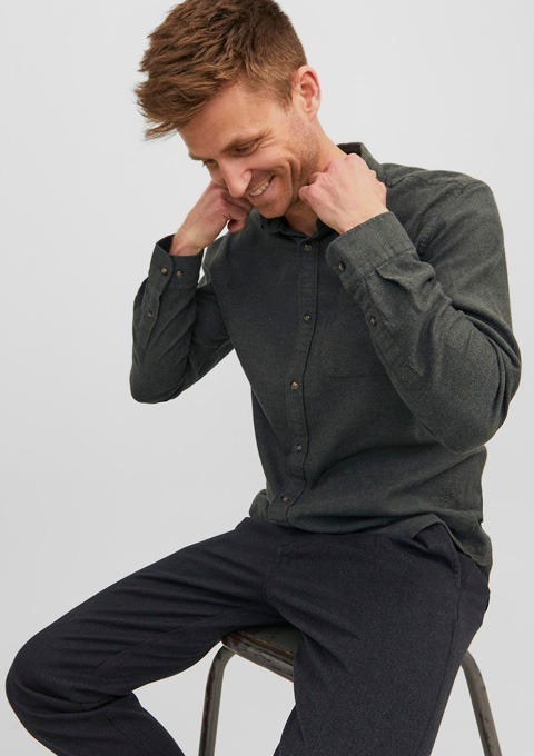 Ein Mann sitzt auf einem Stuhl und zieht sein Hemd hoch, dabei lächelt er. Er trägt ein dunkles, langärmliges Hemd und eine dunkelgraue Hose. Ein Mann sitzt auf einem Stuhl und zieht sein Hemd hoch, dabei lächelt er. Er trägt ein dunkles, langärmliges Hemd und eine dunkelgraue Hose.