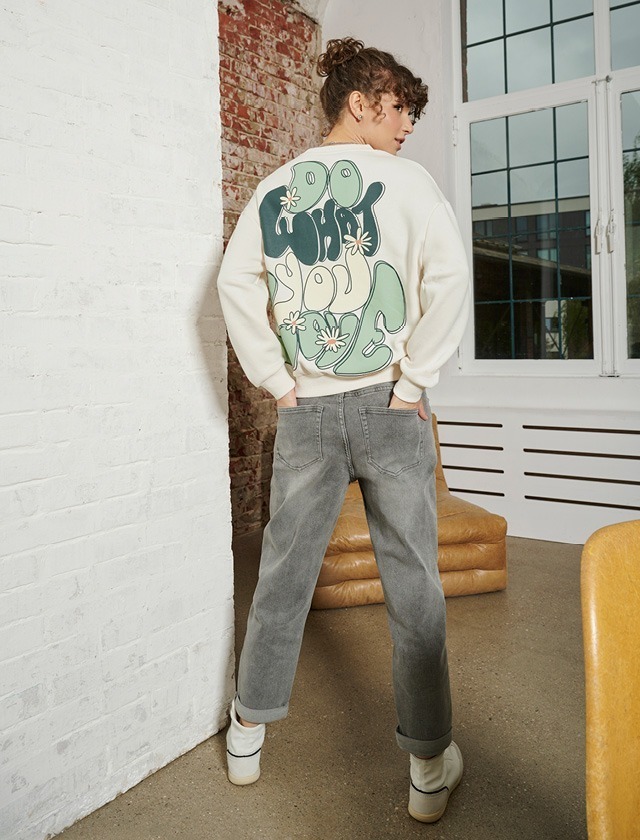 Ein Mädchen mit lockigem Haar trägt einen weißen Pullover mit Aufdruck und graue Jeans. Sie steht mit dem Rücken zur Kamera. Ein Mädchen mit lockigem Haar trägt einen weißen Pullover mit Aufdruck und graue Jeans. Sie steht mit dem Rücken zur Kamera.