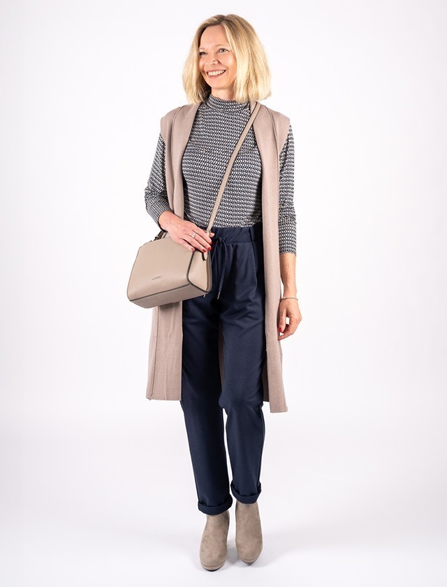 Eine lächelnde Frau trägt einen gestreiften Pullover, eine lange Weste und eine schwarze Hose. Sie hält eine beige Tasche und trägt hellbeige Schuhe. Eine lächelnde Frau trägt einen gestreiften Pullover, eine lange Weste und eine schwarze Hose. Sie hält eine beige Tasche und trägt hellbeige Schuhe.