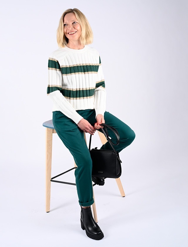 Eine lächelnde Frau sitzt auf einem Hocker, trägt einen gestreiften Pullover und grüne Hose, und hält eine schwarze Tasche. Eine lächelnde Frau sitzt auf einem Hocker, trägt einen gestreiften Pullover und grüne Hose, und hält eine schwarze Tasche.
